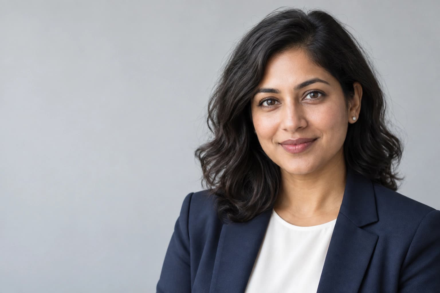 Fictional adult woman with dark wavy hair, silver earrings, navy blazer and ivory shirt against a light gray background.