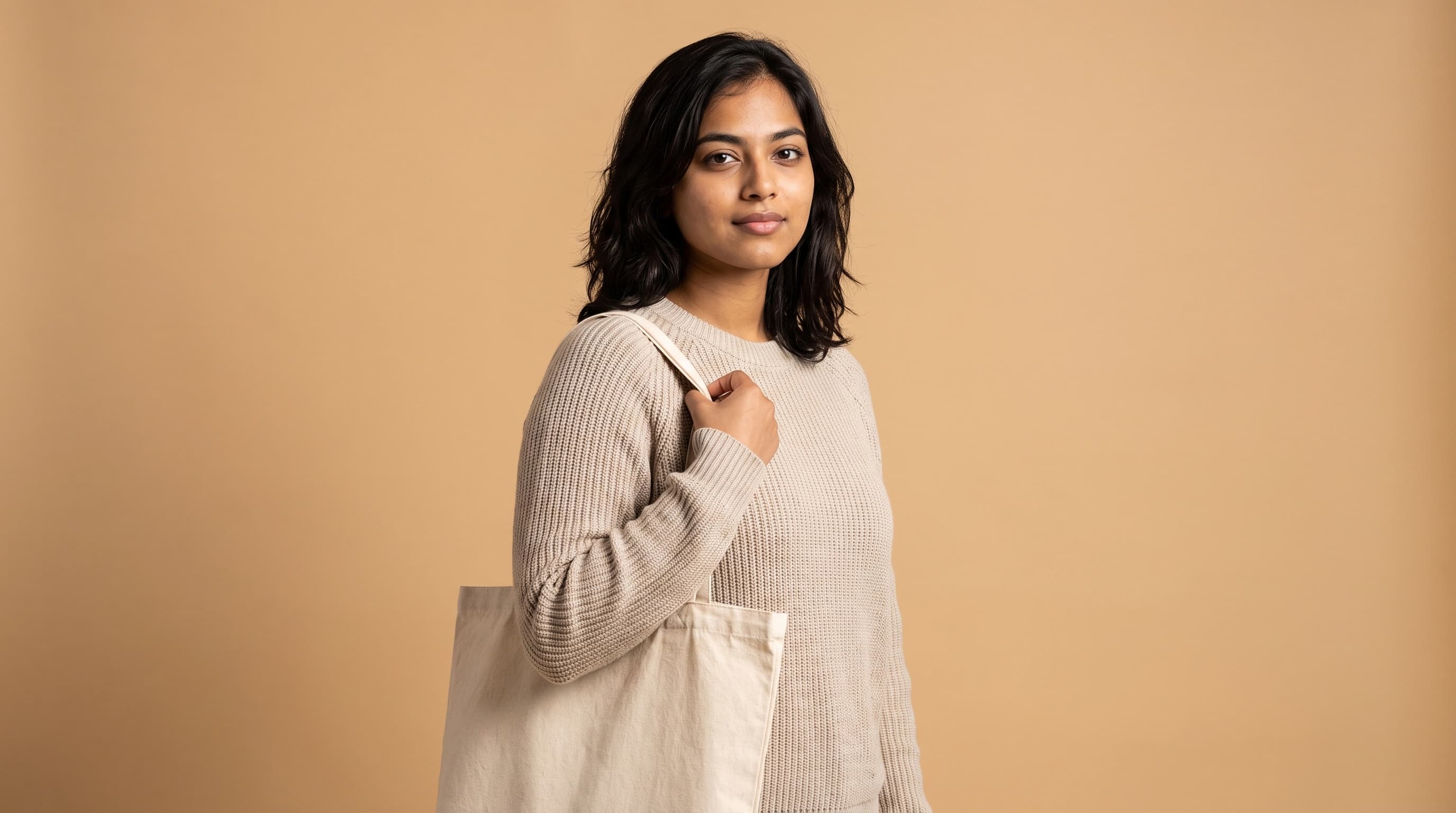 The same synthetic persona photographed against a plain studio backdrop in a plain knit sweater, holding an unbranded canvas tote
