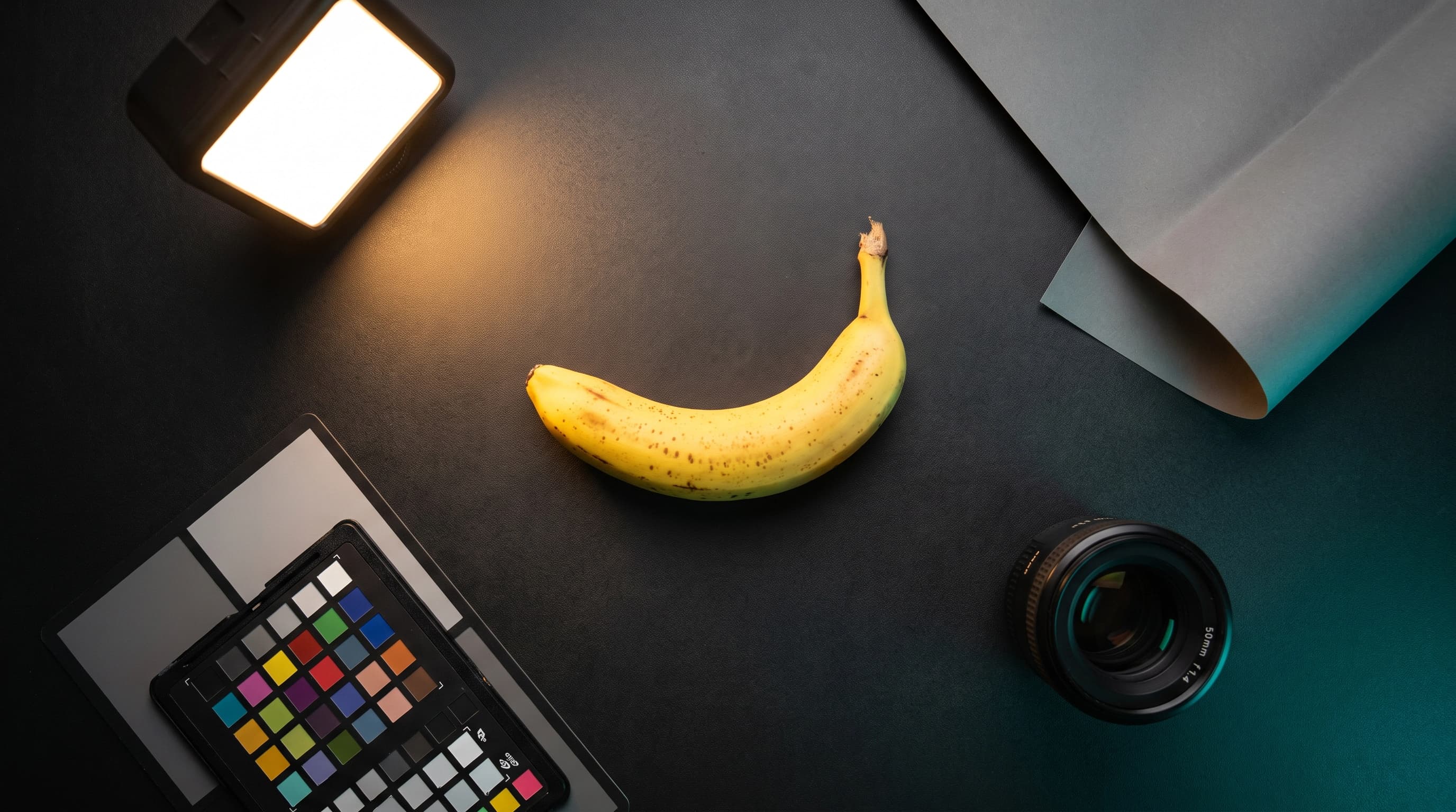 Overhead flat lay of a creative director's desk with a glowing studio lightbox, a color checker chart, a prime lens, and a single ripe banana lit like a hero product
