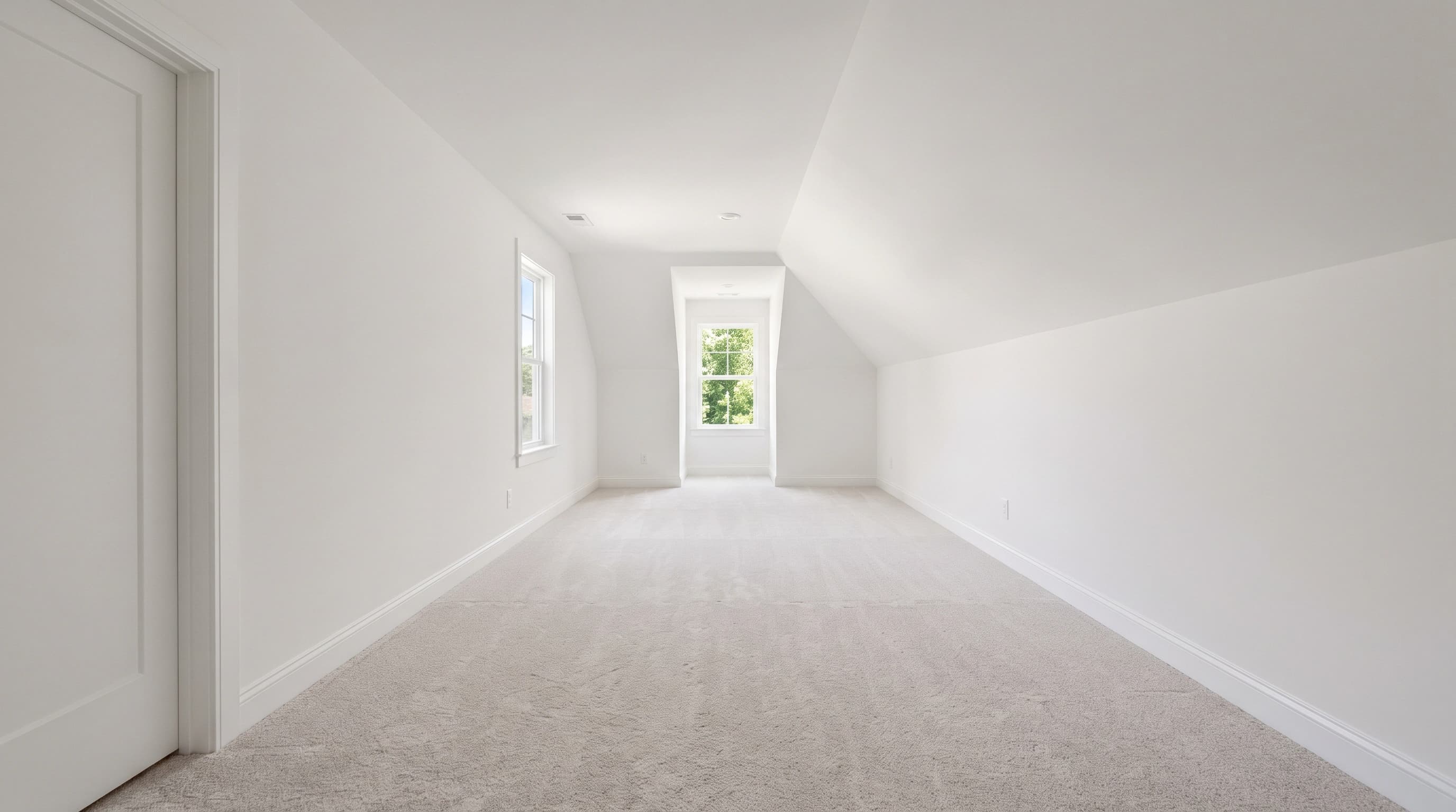Empty long narrow bonus room over a garage with a sloped ceiling, pale carpet, a dormer window at the far end and a small side window, no built-ins or closets