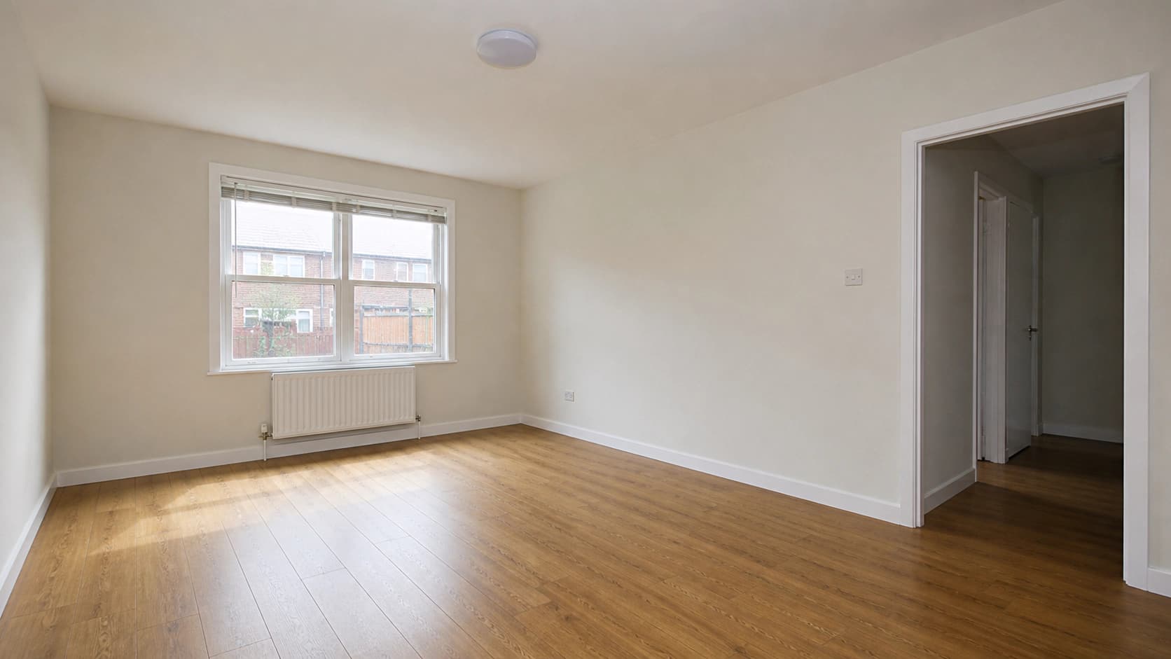 An empty living room photographed from the corner in flat daylight, with a double-hung window and white radiator on the left wall, an open doorway to a hallway on the right, and medium oak laminate flooring