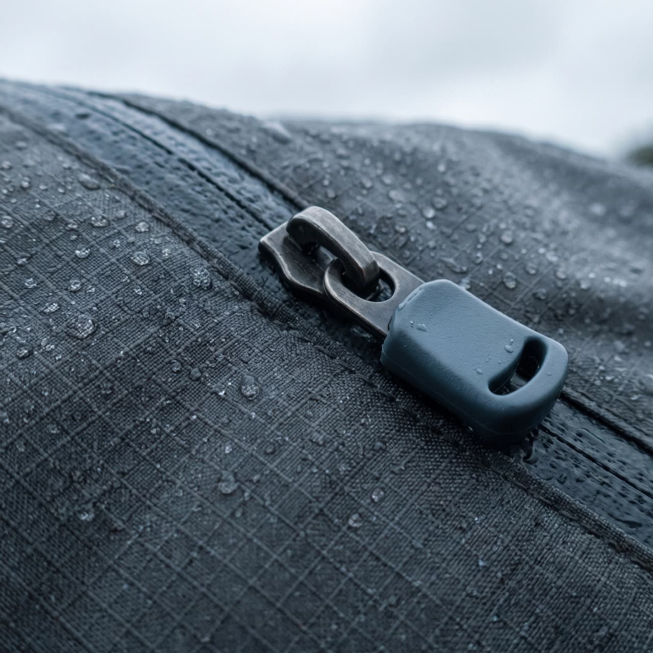 Moodboard tile: macro of graphite ripstop nylon with a brushed steel pull tab and water beads